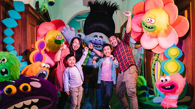 A family of four surrounded by large colourful smiling flowers, a troll and other characters.
