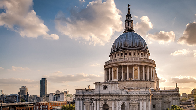 The exterior of St Paul's Cathedral, with its iconic grey-coloured dome, against a bright sky with the sun shining through clouds.
