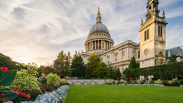 St Paul's Cathedral on a bright day, with wispy cloud in the sky, and a green lawn and colourful flower bed in the foreground.