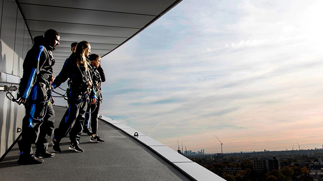 A small group of people, attached to a railing, peer over the edge of Tottenham Hotspur Stadium at sunset, with London's skyline in the background.