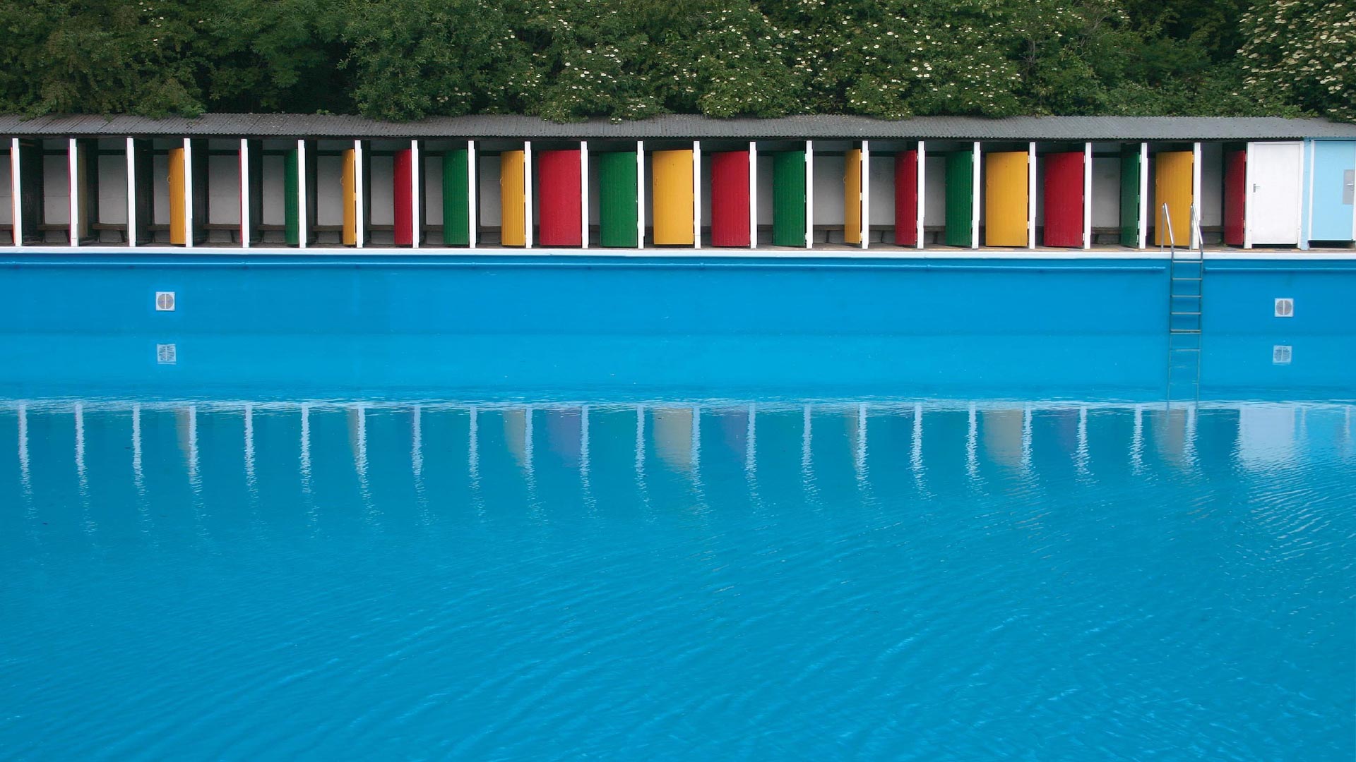 Go for a dip in Tooting Bec Lido. Image courtesy of Tooting Bec Lido. Looking across the pool at the colourful yellow, green and red doors of Tooting Bec Lido.