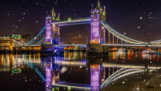 Die Tower Bridge beleuchtet mit lila Lichtern bei Schneefall und die Brücke wird auf die Themse darunter reflektiert. 