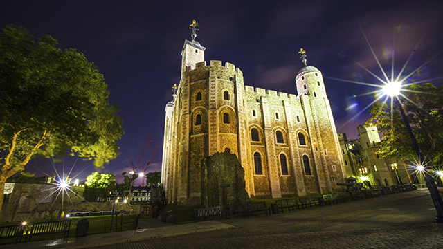 The Tower of London, illuminated in a yellow light, at night. A dimly lit tree on the left, lamp post on the right and a purplish dark sky create a spooky feel.