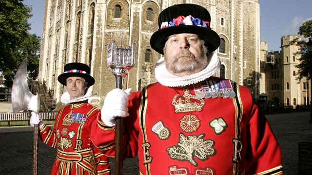 Two yoeman warders dress in red and gold tunics with a black hat stand in front of the tower of london.
