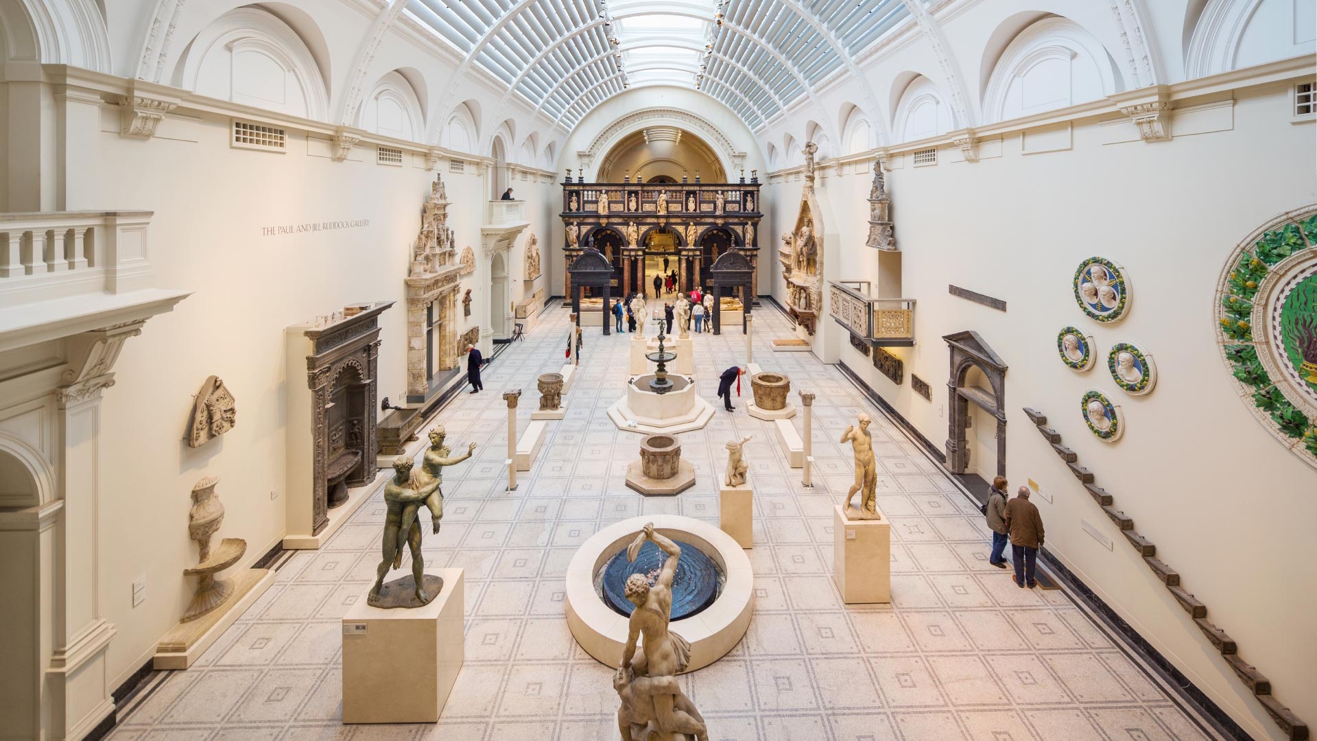 A vast light-filled white hall at Victoria and Albert Museum, featuring  sculptures and statues on pedestals, artefacts on walls and a curved, glass roof.