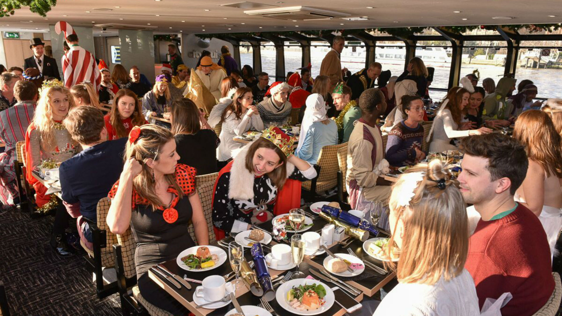 Christmas Day traditional lunch cruise on the river Thames. Image courtesy of City Cruises. A crowd of people wearing festive costumes enjoying a Christmas Day lunch aboard a cruise.