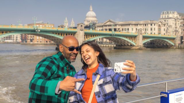 Two people take a selfie next to London bridge on a City Cruises river thames boat tour. 