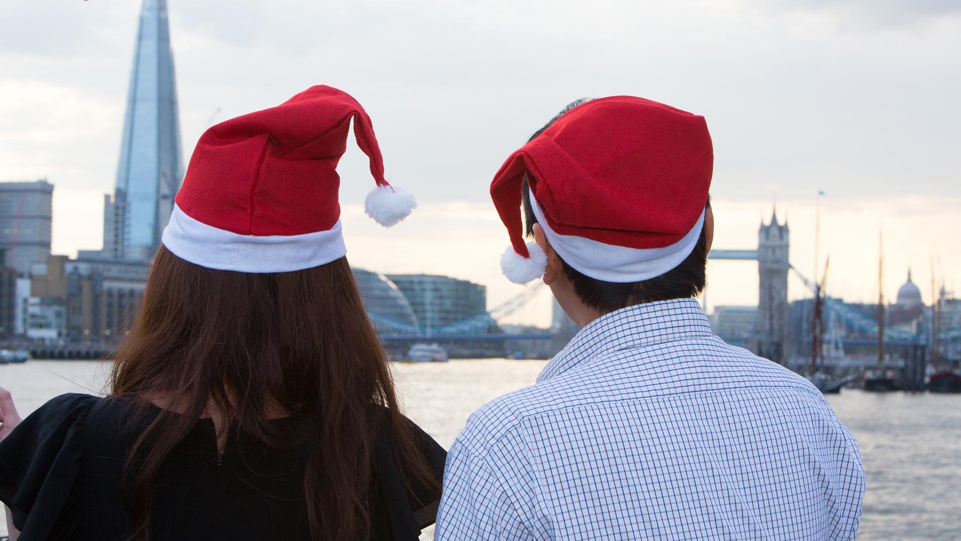 Two people in red Santa hats gaze out at the river Thames, the Shard and Tower Bridge from aboard one of City Cruises' boats.