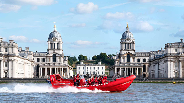 La vedette rouge Thames Rockets, avec ses passagers vêtus de gilets de sauvetage rouges, file sur la Tamise par une journée ensoleillée, avec les dômes jumeaux du Royal Naval College de Greenwich en arrière-plan.