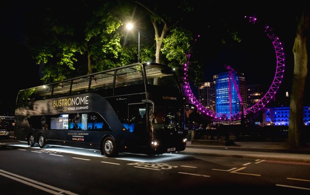 A large black dinner bus with a glass roof parked next to the london eye lit up with pink lights.