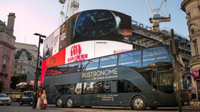 A large black bus with a glass ceiling parked against the large screens in picadilly circus london.