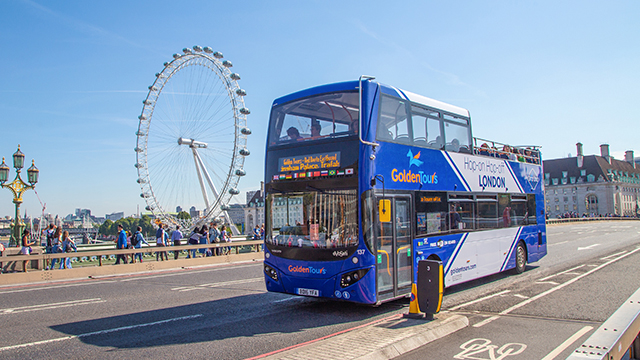 A blue and white Golden Tours double-decker bus passes in front of the London Eye on a cloudless day.