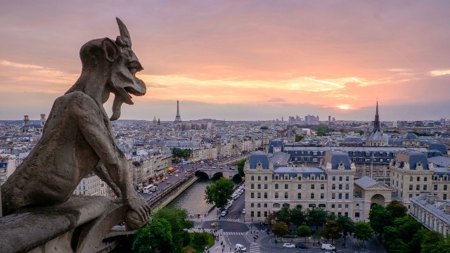 Paris Notre Dame cathedral gargoyle in the foreground overlooking the city with the Eiffel Tower in the distance as the sun sets.
