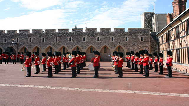 Windsor Castle guardsmen in traduitional attire, red jackets and bear skin hats, are standing in rows in Windsor Castle courtyard. 