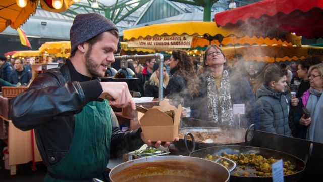 A man in a green apron serves street food from a large pan into a box for customers in borough market. 