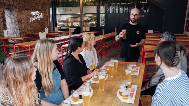 A group of people sit at a long table trying different beers with a guide at a london brewery.