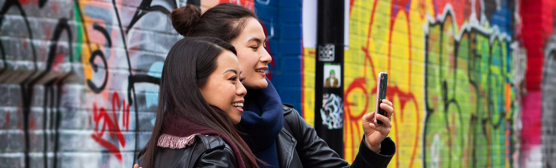 Two women take a selfie in front of a wall covered in colourful street art.