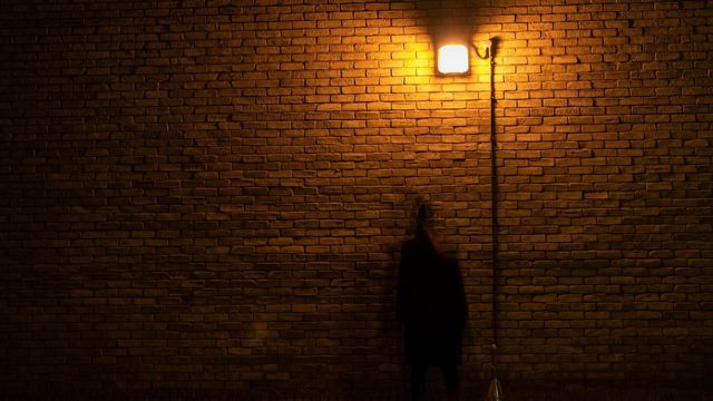 A ghostly figure stands under a street lamp against a brick wall.