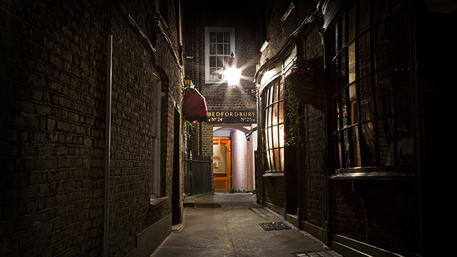 A narrow, darkened alley leads to a dimly lit pub at night, which is part of the London Grim Reaper Tour.