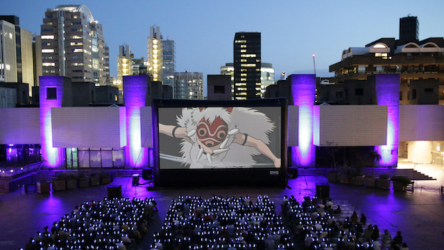 A group of people watch a film on a big screen with the skyscrapers of London in the background at dusk.