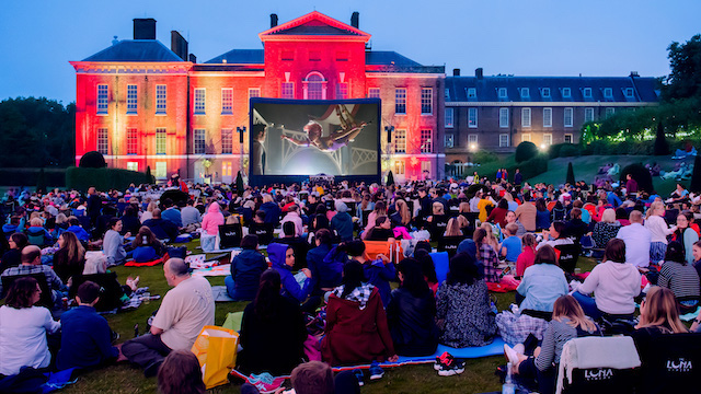 People watch a film on a big screen outside Kensington Palace in London at dusk.