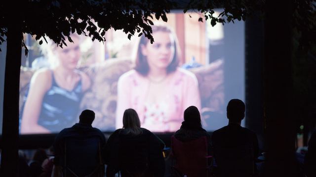 People in silhouette watch a film on a large outdoor screen in London at dusk.