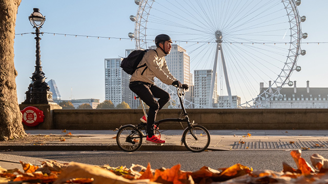 A man cycling on a bridge with the London Eye in the background and autumn leaves in the foreground.