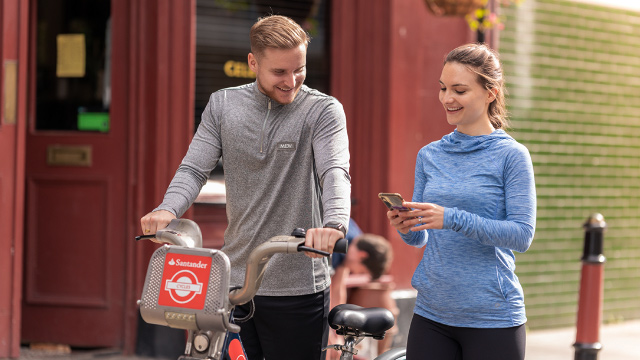 A man on a Santander Cycle stands next to a woman on a smartphone in London