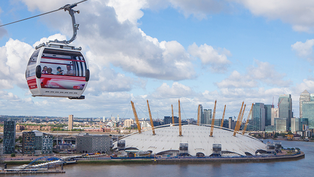One of the cabins on the London cable car moves in the air, with The O2 and river Thames in the background, on a slightly cloudy day.