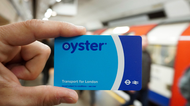 Person holding a blue London transport Oyster card with a London Underground train arriving at a Tube platform in the background.