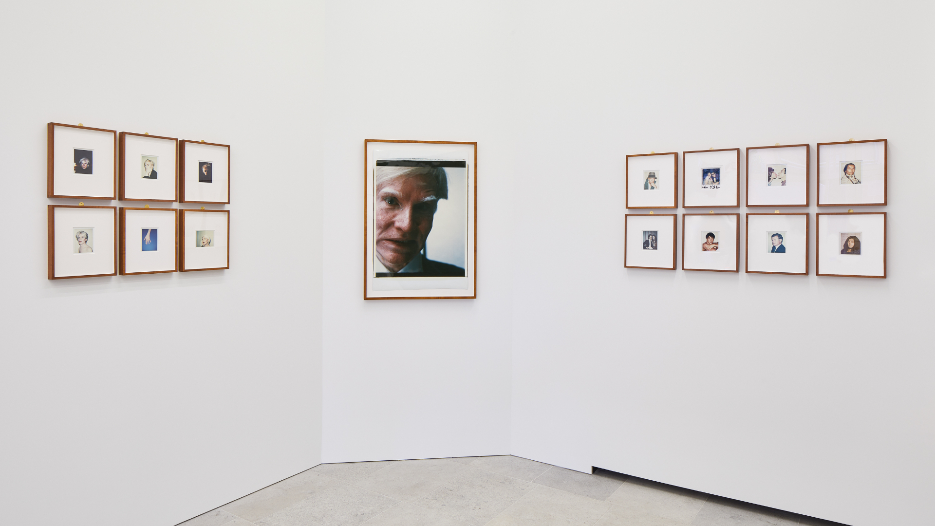 A collection of framed Polaroid photographs on a white wall, including a large self-portrait, by Andy Warhol as part of Andy Warhol Polaroid Pictures exhibition at Bastian