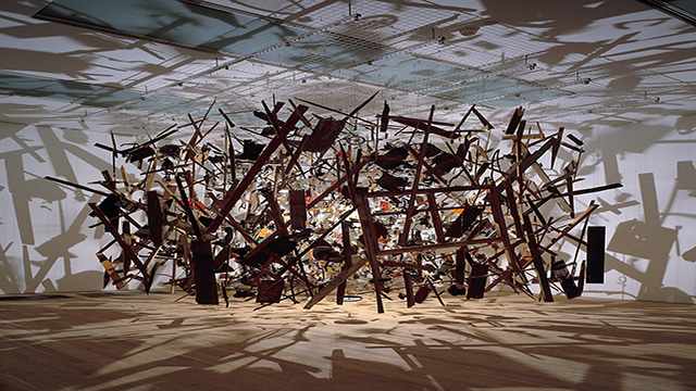 Exploded parts of a garden shed suspended in the air, with the light reflecting the shades of the different parts of the sculpture.