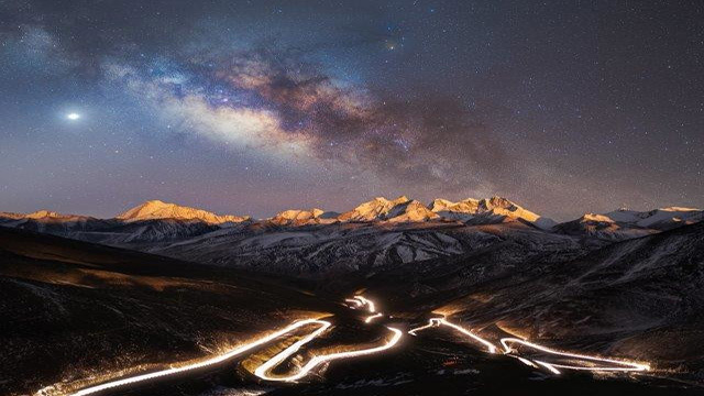The Milky Way illuminates the landscape almost reflecting the illuminated National Highway 219, with mountains on the background.