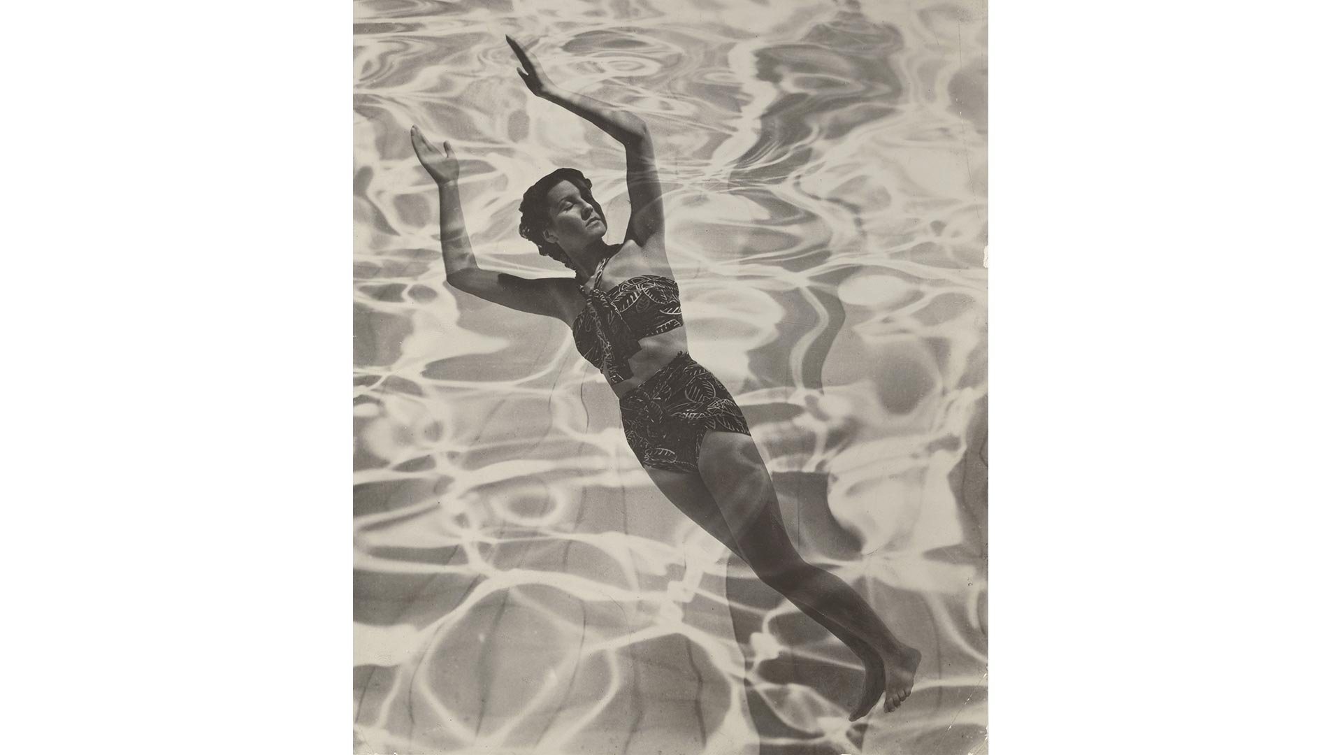 A model floats outstretched in water, which is dappled with sunlight, in a black-and-white photograph.