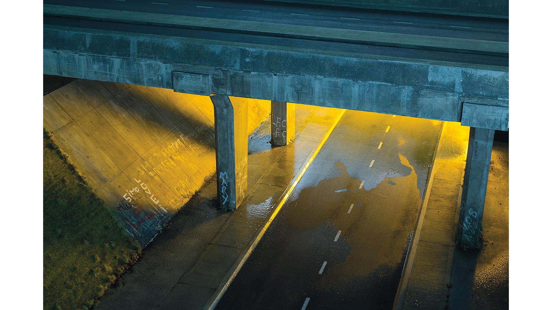 A road crossing beneath a motorway bridge, illuminated at night, with graffiti on the walls.