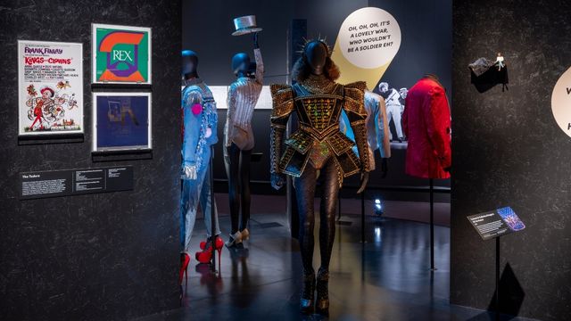 A gold sparkling costume bodysuit with high shoulder pads against a backdrop of posters and images from SIX the musical at reimagining musical exhibition at the V&A