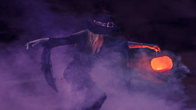 Une femme, déguisée en sorcière, semble dancer dans un brouillard de couleur violette, tout en tenant une citrouille illuminée.