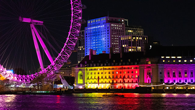 Das London Eye ist nachts violett beleuchtet, und die Gebäude am Flussufer leuchten in Regenbogenfarben. 