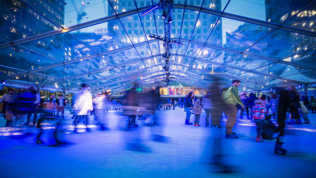 Ice skaters skate around the ice rink at canary wharf in london, lit in blue light and surrounded by skyscrapers.