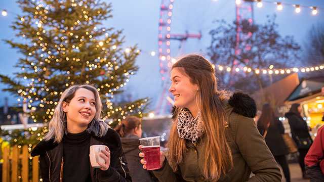 Zwei Mädchen genießen abendliche Drinks auf einem Londoner Weihnachtsmarkt mit einem Weihnachtsbaum, Riesenrad, Marktständen und Lichtern im Hintergrund 