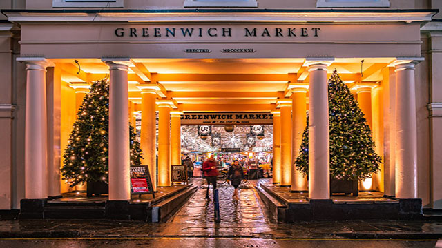 Eingang zum Greenwich Market mit Säulen vor geschmückten Weihnachtsbäumen und vorbeilaufenden Menschen.