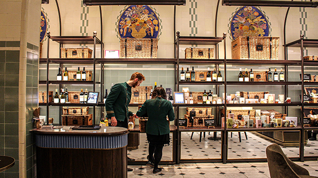 Two staff stand in a shop in Harrods, wearing the iconic green uniform with gold buttons, looking at items to place on the shelves. 