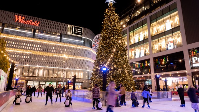 A giant traditional Christmas tree, covered in warm white fairy lights and a big star on top, in the centre of the ice rink filled with people and in front of Westfield Shopping Centre.