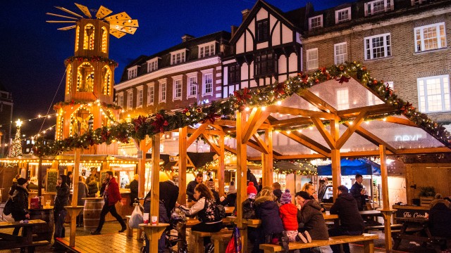Menschen sitzen, essen und trinken unter einem gut beleuchteten, mit Weihnachtsgirlanden, Lichtern und Ornamenten geschmückten Marktplatzdach in einer historischen Marktstadt.