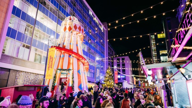 Fairy lights strung from building to building as crowds of people walk around outside the well lit Selfridges Christmas market.