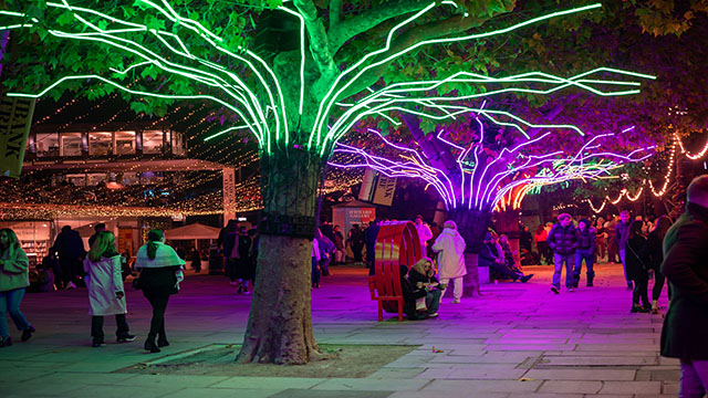 Gruppen von Menschen spazieren an Holzhütten des Southbank-Weihnachtsmarktes vorbei, umgeben von neonbeleuchteten Bäumen.