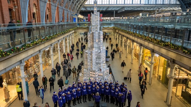 A choir of people singing in front of the St Pancras station Christmas tree, made from white paper and illustrated with drawings of Christmas symbols and the London skyline.