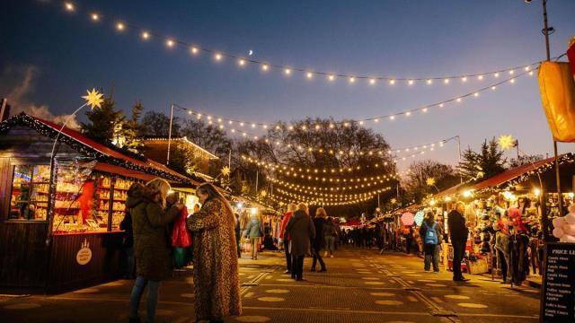 Personen mit Mänteln besuchen Weihnachtsmarkt Hütten auf beiden Seiten, mit hängenden funkelnden Lichterketten in der Dämmerung.