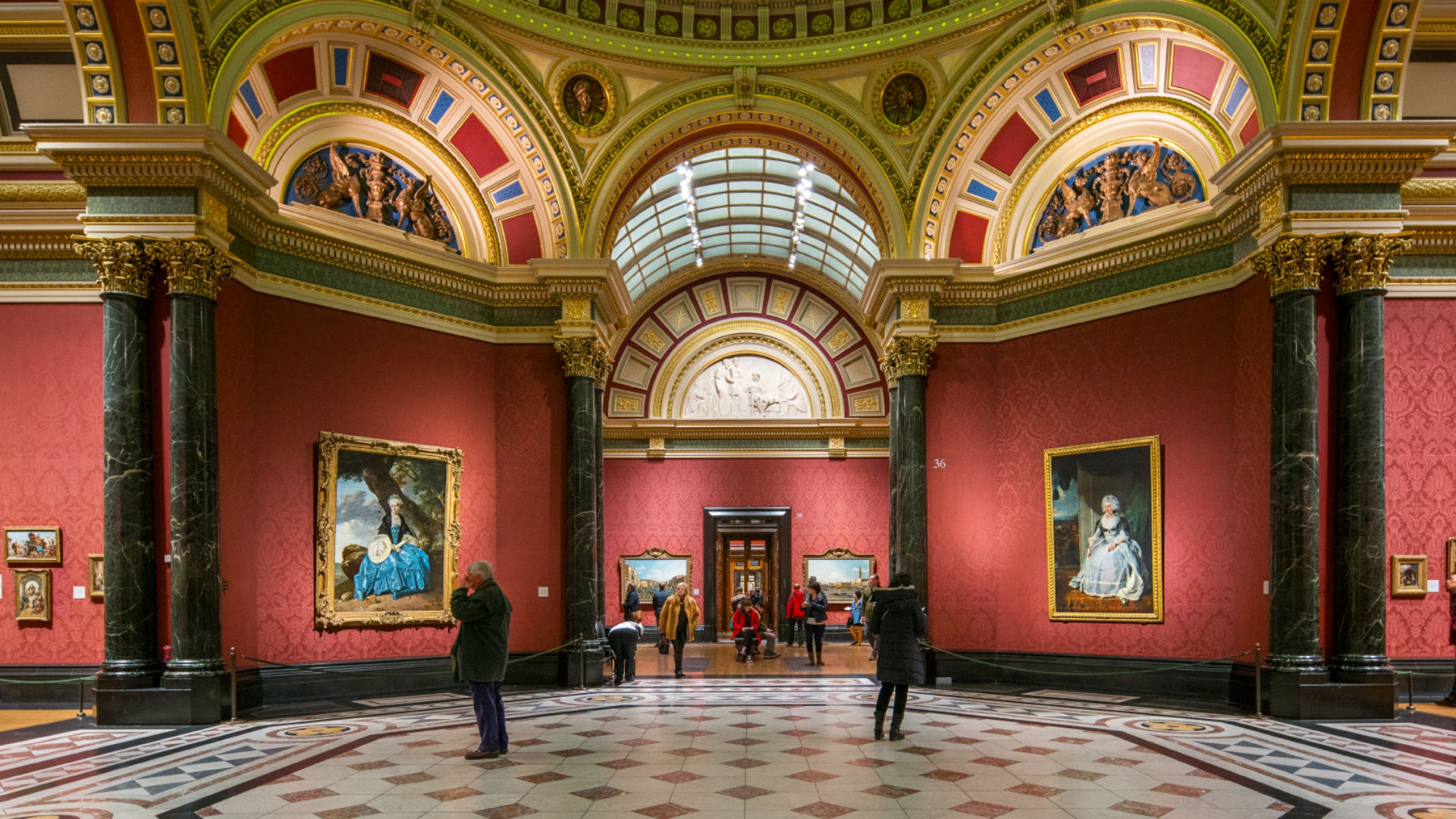 Two people looking at paintings in the red-painted halls of the National Gallery.
