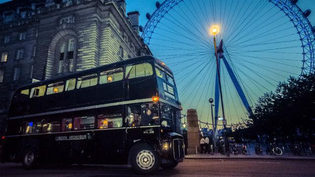 A photo of a black Ghost Bus driving London's streets, with the Coca-Cola London Eye in the background.